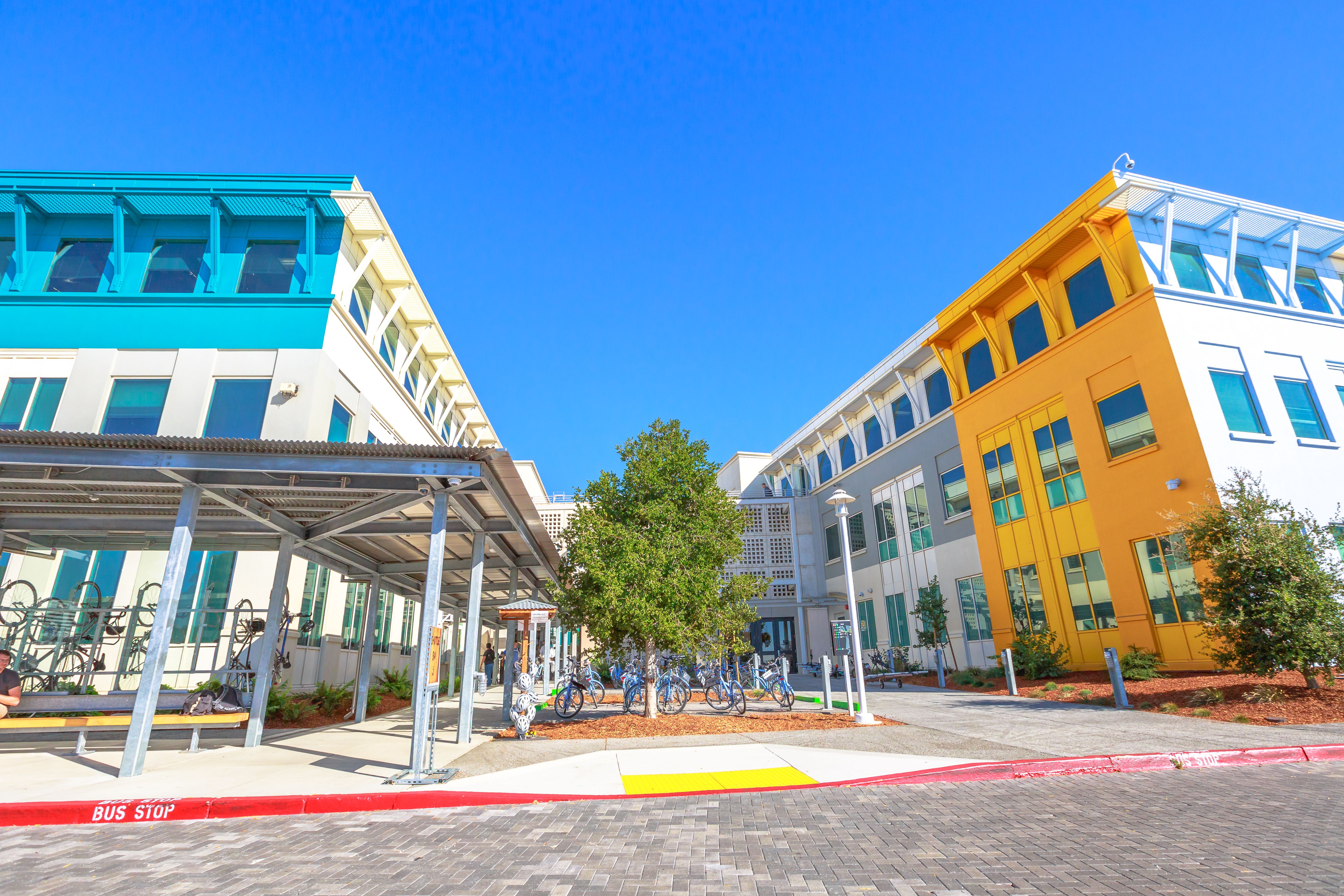 The Facebook HQ building--there is an orange/yellow block on the right side of the building and a blue one on the left. There are bikes locked and a bus stop